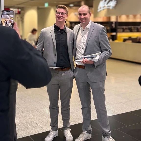 Ein Bild von zwei lächelnden Männern in grauen Anzügen die sich von einem weiteren Mann mit einem Handy fotografieren lassen