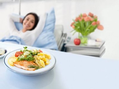 Frau liegt im Krankenhausbett lächelnd und schaut auf ein Tellergericht