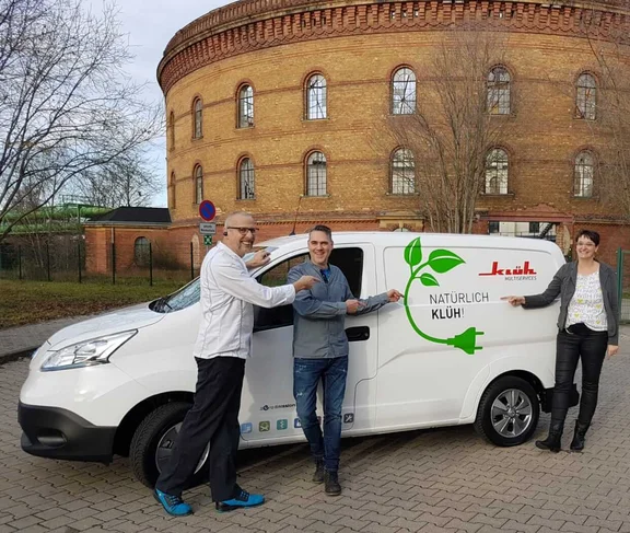drei lächelnde Personen die neben einem Klüh Multiservice Firmenwagen stehen und auf den Schriftzug "Natürlich Klüh!" auf dem Auto zeigen