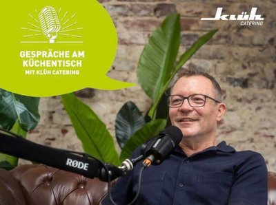 Bild vom Thorsten Greth sitzend auf einer Couch mit einem Mikrofon vor sich zum Klüh Catering Podcast "Gespräche am Küchentisch"
