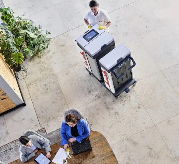 in einem großen Raum ist eine Klüh Reinigungskraft stehend an ihrem Reinigungswagen und zwei Büroangestelle Frauen, die an einem Tisch sitzen und sich besprechen