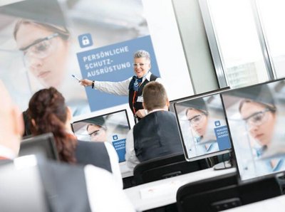 Ein Bild von einem Seminarraum mit vielen Computern an dem Menschen sitzen und nach vorne zur Leinwand schauen, wo eine lächelnde Frau steht und auf die Leinwand zeigt wo ein Bild präsentiert ist mit dem Titel " Persönliche Schutzausrüstung"