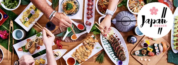 Aktionsbild zur "Japan Wochen" mit einem Tisch voller japanischen Speisen und Hände mit Ess Stäbchen die danach greifen