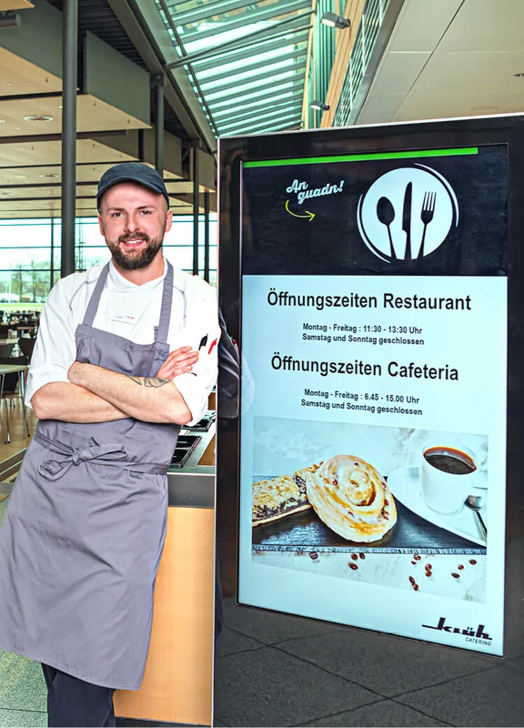 Ein Bild von einem lächelnden Klüh Catering Koch, der seine Arme verschränkt und sich an ein Digitale Werbetafel lehnt auf dem die Öffnungszeiten eines Restaurants & Cafateria stehen