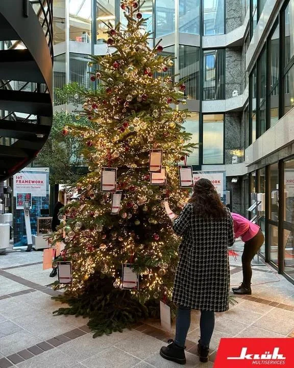 ein beschmückter Weihnachtsbaum an dem Wunschzettel hängen. Um dem Baum stehen Menschen.