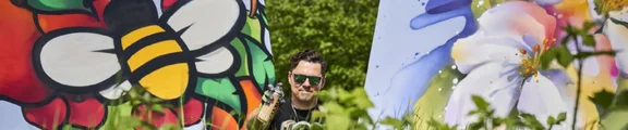 Künstler Damian Bautsch mit einer Graffiti-Dose in der Hand zwischen zwei großen Gemälden