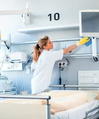 Eine Frau in weißer Kleidung reinigt die Ablage oberhalb eines Krankenhausbettes. Im Hintergrund sind Bildschirme und medizinische Technik zu erkennen.
