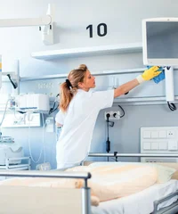 Eine Frau in weißer Kleidung reinigt die Ablage oberhalb eines Krankenhausbettes. Im Hintergrund sind Bildschirme und medizinische Technik zu erkennen.