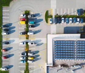 Ein Bild aus der Vogelperspektive von einem Gelände mit einem Gebäude mit Solaranlagen auf dem Dach und ein Parkplatz auf dem viele Autos parken 