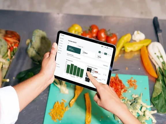 Ein Bild von einer Person in der Küche, auf dem Tisch liegen viele Gemüse und Obst Sorten sowie ein Schneidebrett und Messer. Die Person hält eiin Tablet in der Hand und sieht sich eine Auswertung an.