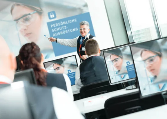 Ein Bild in einem Raum mit einigen Menschen die in Reihen hintereinander sitzen am PC und schauen nach vorne, wo eine Frau eine Schulung hält mit dem Titel: Persönliche Schutzausrüstung