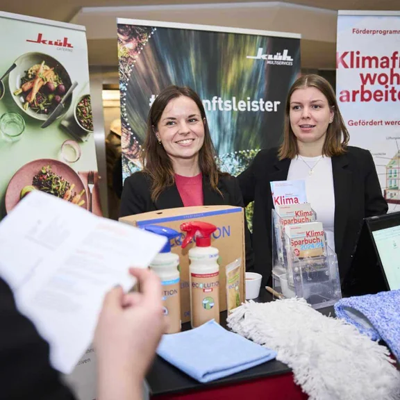 Infostand von Klimamobil Düsseldorf und Klüh Multiservices, zwei Frauen stehen lächelnd hinter dem Stand und betreuen Besucher