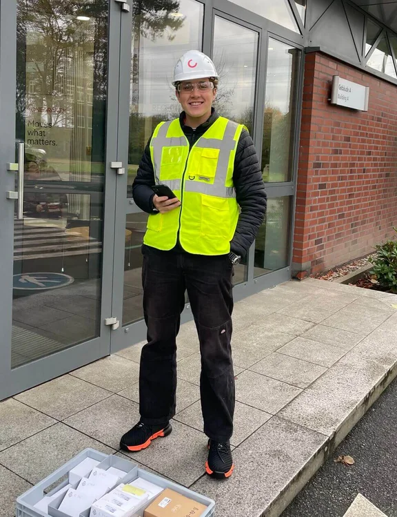 junger Mann in Schutzarbeitskleidung mit Helm und Warnweste lächelt in die Kamera, vor ihm eine graue Kiste mit kleinen Paketen