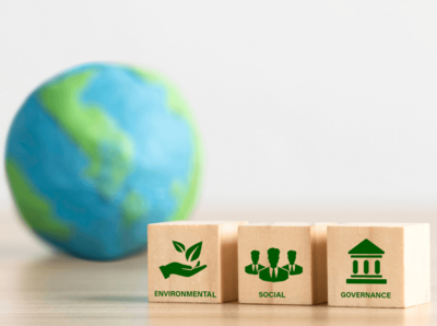 im Vordergrund 3 Holzwürfel, auf dem ersten Würfel ist ein grüner Schriftzug Environmental mit grünem Logo einer offenen Hand welche Laubblätter fängt, auf dem zweiten Würfel ist der schriftzug Social und als Logo drei Umrisse von Menschen, auf dem dritten Würfel ist der Schriftzug Governance mit dem Logo eines Regierungsgebäudes. Im Hintergrund ist leicht verschwommen eine Globus zusehen.