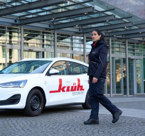 Eine weibliche Sicherheitskraft passiert ein weißes Auto mit Klüh-Logo.
