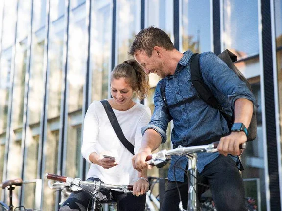 ein Mann und eine Frau auf ihren Fahrrädern die lächeln und dabei auf ein Smartphone schauen