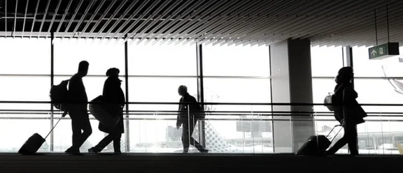 Bannerbild im Gebäude eines Flughafens, einige Menschen mit Koffern und Taschen gehen durchs Bild
