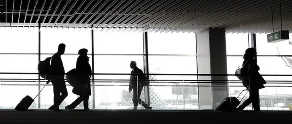 Bannerbild im Gebäude eines Flughafens, einige Menschen mit Koffern und Taschen gehen durchs Bild
