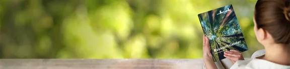 Ein Bild von einer Frau die eine Zeitschrift ließt mit dem Titel #zukunftsleister Nachhaltigkeitsbericht 2023 von Klüh Multiservices" im Hintergrund ist grüne Naturlandschaft