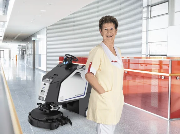 Klüh Cleaning Angestellte schaut lächelnd und steht vor einer Scheuersaugmaschine