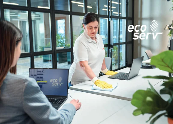 Eine Frau am Schreibtisch arbeitet an ihrem Notebook und eine Klüh Cleaning Reinigungskraft wischt ein beistehenden Tisch mit einem blauen Tuch