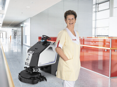 Klüh Cleaning Angestellte steht lächelnd in Arbeitskleidung vor einem Reinigungsroboter in einem Gebäude
