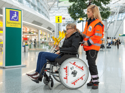 Ein Bild von einen Innenbereich des Flughafens. Im Vordergrund sitzt eine Frau im Rollstuhl mit der beschriftung Klüh Airport Service und wird angeschoben von einer Frau in Arbeitskleidung